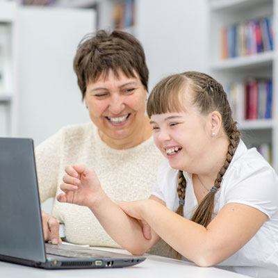 Smiling girl with down syndrome uses a laptop with her teacher in the library